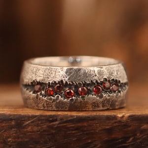 May include: A silver ring with a textured finish, featuring a line of small, red gemstones. The ring is set against a blurred, warm-toned backdrop, accentuating the contrast between the metal and the gems.