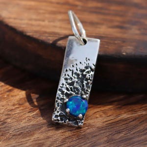 Puede incluir: Un colgante de plata con una superficie texturizada y una piedra de ópalo azul.