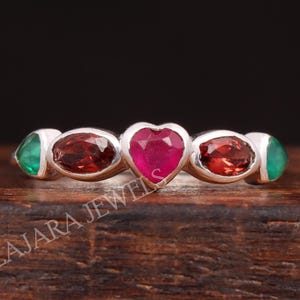 May include: A silver ring featuring a heart-shaped ruby center stone, flanked by oval-cut garnet and emerald gemstones. The ring is set against a dark background, highlighting the vibrant colors of the gemstones.