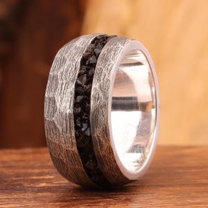 May include: A silver ring with a textured band and a central inlay of small, dark gemstones. The ring is set against a wooden surface, showcasing its design and craftsmanship. The ring's band has a hammered texture.