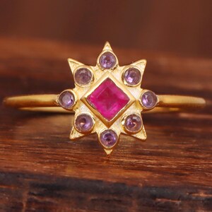 Könnte beinhalten: Ein goldfarbener Ring mit Sternmuster. In der Mitte befindet sich ein quadratischer, leuchtend rosa Edelstein, umgeben von kleineren, runden, lila Edelsteinen. Der Ring liegt auf einer Holzoberfläche.