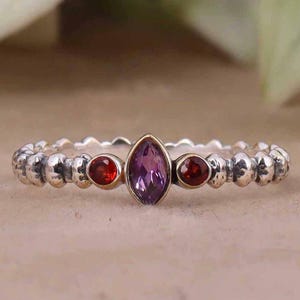 May include: A silver ring with a purple amethyst gemstone and two red garnet gemstones. The ring has a beaded band.