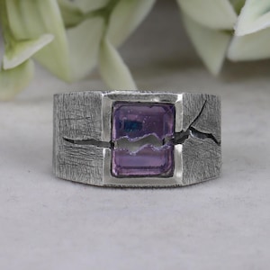 May include: A silver ring with a textured band and a square-shaped purple gemstone set in the centre. The band has a crack running through it, creating a unique design.