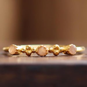 Könnte beinhalten: Ein goldener Ring mit drei kleinen, hellrosa Steinen, die in einem strukturierten Band gefasst sind.