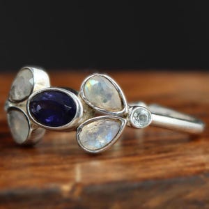 Puede incluir: Un anillo de plata con un grupo de cinco piedras preciosas. El anillo presenta una gran amatista ovalada rodeada de cuatro piedras de luna en forma de pera.