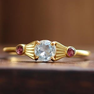 以下が含まれることがあります： 淡いブルーの宝石と、両側に小さなピンクの宝石が2つ付いたゴールドのリングです。