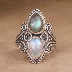 Puede incluir: Un anillo de plata con una piedra de luna en forma de pera y una piedra de labradorita en forma de pera. El anillo tiene un diseño detallado y adornado.