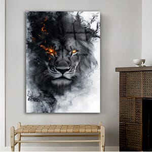 May include: A framed art print featuring a lion's face with a dark, smoky mane and fiery orange eyes. The artwork is predominantly black and white, with the lion's face centered and the smoke-like elements surrounding it. The print is displayed on a white wall.