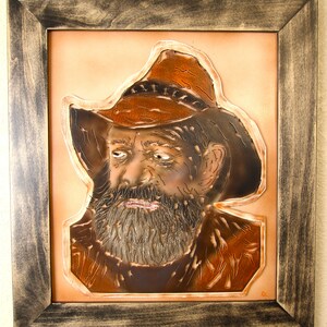 May include: A leather portrait of a man with a beard wearing a brown cowboy hat. The portrait is framed in a dark wood frame.