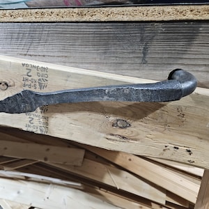 Może przedstawiać: Kute metalowe narzędzie z zakrzywionym uchwytem i spiczastym końcem. Narzędzie jest ciemnoszare i spoczywa na drewnianej powierzchni. Drewno ma widoczne słoje i sęki. Wygląda na rodzaj dźwigni lub łomu.