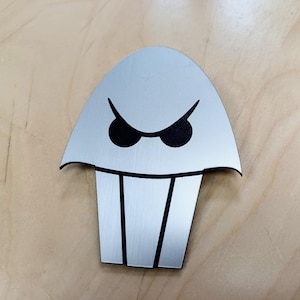 May include: A silver metal emblem featuring a cartoon ghost design. The ghost has black eyes and a wide mouth with two black vertical lines. The emblem is on a light brown wooden surface.