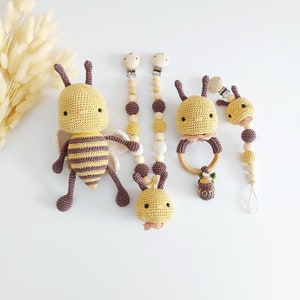 May include: A collection of handmade crochet baby toys in a bee theme. The set includes a large bee plush, two pacifier clips with wooden beads, a wooden ring teether, and a smaller bee toy. The toys are yellow, purple, and beige.