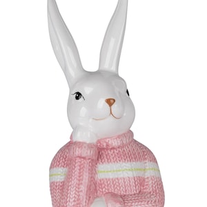 Könnte beinhalten: Eine weiße Keramik-Hasenfigur mit langen Ohren und einem rosa Pullover. Der Pullover hat weiße und hellgrüne Streifen. Der Hase hat eine braune Nase und stützt sein Kinn auf seine Pfote. Ein dekorativer Artikel zum Thema Ostern oder Frühling.