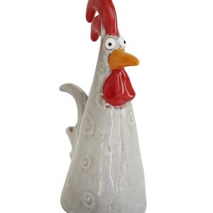 Könnte beinhalten: Eine skurrile Keramik-Hahnfigur. Der Hahn hat einen grauen Körper, einen roten Kamm und Kehllappen sowie einen orangefarbenen Schnabel. Die Figur hat einen überraschten Gesichtsausdruck und ist mit kleinen, kreisförmigen Vertiefungen verziert.