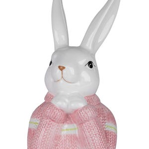 Könnte beinhalten: Eine weiße Keramik-Hasenfigur mit langen Ohren und einem rosa Pullover. Der Hase hat schwarze Augen und eine braune Nase und hält die Pfoten zusammen. Der Pullover hat weiße und gelbe Streifen.