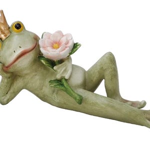 Könnte beinhalten: Eine skurrile Froschfigur, die sich zurücklehnt und eine rosa Blume hält. Der Frosch ist hellgrün mit gelben Augen und einer goldenen Krone. Ein dekorativer Artikel für Haus oder Garten.