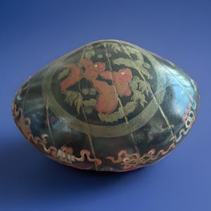 May include: A black, oval-shaped decorative object with a floral design in red and gold. The object has a glossy finish and is set against a blue background. The floral design is centered on the top.