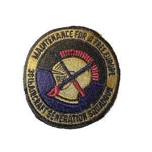 May include: A circular embroidered patch with a gold background and black border. The patch features a blue and gold helmet with crossed swords and the text "Maintenance for a Free Europe" and "Seacat Generator Squadron".