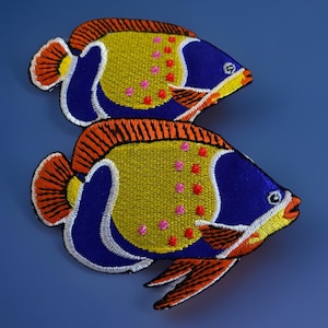 May include: Two embroidered fish patches. Each patch features a blue head, yellow body with pink and red dots, and orange fins. The fish are outlined in white and set against a blue background. These patches could be used to decorate clothing or accessories.