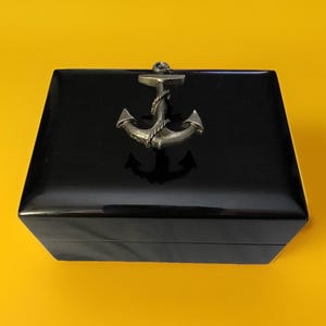 May include: A black, rectangular box with a shiny finish. A silver-colored anchor is affixed to the top. The anchor is detailed with rope and is centered on the lid. The box is set against a yellow background.