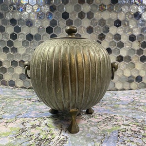 May include: A round, silver-colored metal container with a lid and three legs. The container has a decorative design around the top and is ribbed vertically.