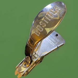 May include: A vintage gold-toned Bostitch staple remover. The word "BOSTITCH" is embossed on the top. The staple remover is open, revealing the silver-colored metal interior and the staple removal mechanism. The background is a solid green.