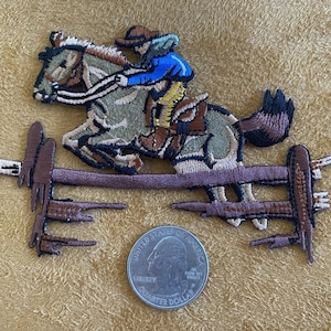 May include: Embroidered patch featuring a horse and rider jumping over a fence. The rider wears a blue shirt and brown hat. The horse is brown and tan. A US quarter coin is next to the patch.