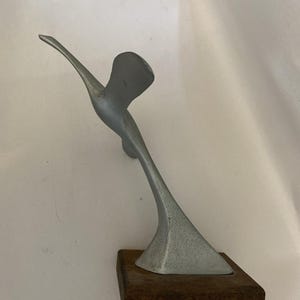 May include: A silver-colored sculpture of a bird in flight, mounted on a dark brown wooden base. The bird has outstretched wings and a streamlined body, creating a sense of motion. The sculpture is likely made of metal.