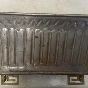 May include: A rectangular metal planter with a weathered, aged appearance. The planter has a flared shape with vertical, rounded grooves and decorative trim along the top edge. It features two decorative, square-shaped feet.