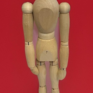 May include: A wooden artist's mannequin with articulated joints, standing on a round base. The figure is made of light-colored wood and is posed against a solid red background. The mannequin is designed for drawing and posing.