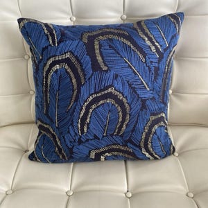 May include: A square decorative pillow with a blue feather pattern. The pillow features a dark blue background with embroidered feathers in shades of blue and gold. The pillow is placed on a white surface.