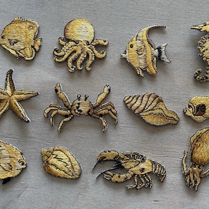 May include: Embroidered patches featuring various sea creatures, including fish, an octopus, a seahorse, a starfish, a crab, a seashell, a lobster, and a squid. The patches are in shades of gold and brown, with detailed stitching.