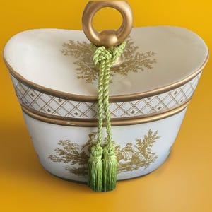 May include: An oval-shaped, off-white ceramic container with a gold handle and trim. The container features a decorative gold pattern and a green tassel. The background is a solid yellow.