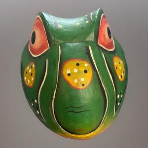 May include: A hand-painted wooden frog mask. The mask is primarily green with yellow and white accents. It features large red and black eyes, and a yellow circle with black and white dots on the forehead. The mask has a whimsical, folk art style.