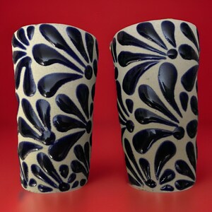 May include: Two ceramic tumblers with a cream-colored base and a repeating pattern of dark blue floral designs. The tumblers have a slightly irregular shape and a glossy finish, set against a red background.