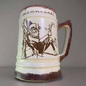 May include: A cream-colored ceramic beer mug featuring a brown speckled rim and base. The front displays a brown graphic illustration of a bearded figure in a hat, holding a tool, with a rifle and jug nearby. The word Tennessee is written in script.