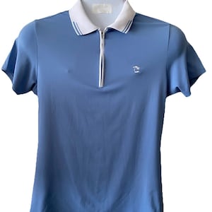 May include: A light blue polo shirt with a white collar and a quarter-length zipper. The collar has two thin blue stripes. A small embroidered emblem is on the left chest. The shirt has short sleeves.