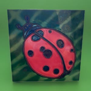 May include: Square ceramic tile featuring a vibrant red ladybug with black spots. The ladybug is set against a dark green background with a leaf-like pattern. The tile has a glossy finish and a decorative appeal.