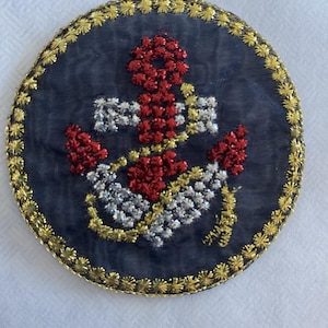 May include: Circular navy blue embroidered patch featuring a red, silver, and gold anchor design. The anchor is the central focus, with intricate stitching details. The patch is bordered with gold floral embroidery.