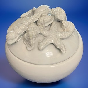 May include: A white ceramic trinket box with a lid, adorned with seashell and starfish details. The round box is painted in a solid white color, with a blue background.