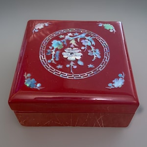 May include: A square, red lacquered box with a decorative floral design inlaid with mother-of-pearl. The lid features a central circular design with flowers and a border, and smaller floral accents at the corners.