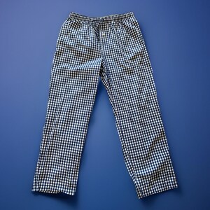 May include: A pair of black and white checkered pajama pants with a drawstring waist. The pants are made of a lightweight fabric and are laid flat on a blue surface. The checkered pattern is evenly spaced.