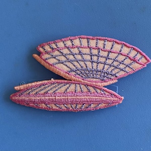 May include: Two embroidered patches in a fan or wing shape. The patches have a light orange background with a dark purple grid pattern. The edges are outlined in a pinkish-red color. The patches are on a blue surface.