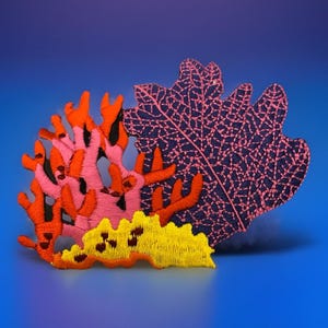 May include: Embroidered coral reef design in shades of pink, orange, and yellow, set against a gradient blue background. The coral features intricate stitching details, creating a textured and vibrant appearance. The design evokes an underwater theme.