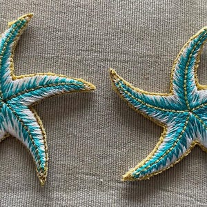 May include: Two embroidered starfish with turquoise and white thread detailing. The starfish have a gold-colored border and are set against a neutral background. These decorative items are likely for coastal or nautical-themed decor.