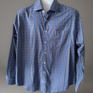 May include: A blue plaid button-down shirt with long sleeves. The shirt features a classic collar, a chest pocket, and white buttons. The plaid pattern includes white, navy, and tan lines. The shirt is displayed on a white mannequin.