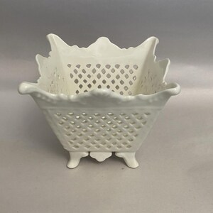 May include: A white ceramic basket with a lattice design and scalloped edges. The square basket sits on three small feet and has a decorative, openwork pattern. The basket is likely for decorative purposes.