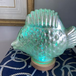 May include: A decorative fish-shaped lamp with a clear, iridescent finish and green glitter accents. The lamp sits on a wooden base and emits a soft, turquoise glow. The lamp is a unique home decor item.