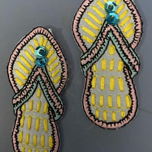 May include: Two embroidered patches in the shape of sandals. The patches have a light gray base with yellow and blue stitching. The edges are outlined in pink and black, and each patch has two blue sequins.