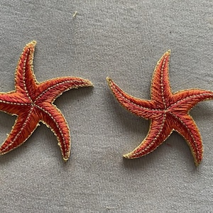May include: Two embroidered starfish patches. Each patch features a five-pointed star shape with a vibrant orange-red hue. The edges are outlined with gold thread, and the details are meticulously stitched.